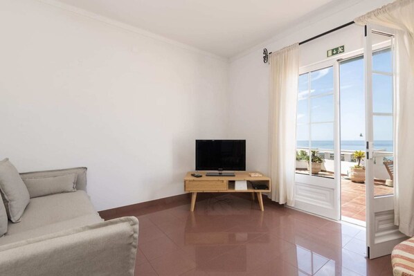 Ein Zimmer mit einer Couch, einem Fernseher auf einem Holzboden, einer Tür zu einem Balkon mit Topfpflanzen und Blick aufs Meer.