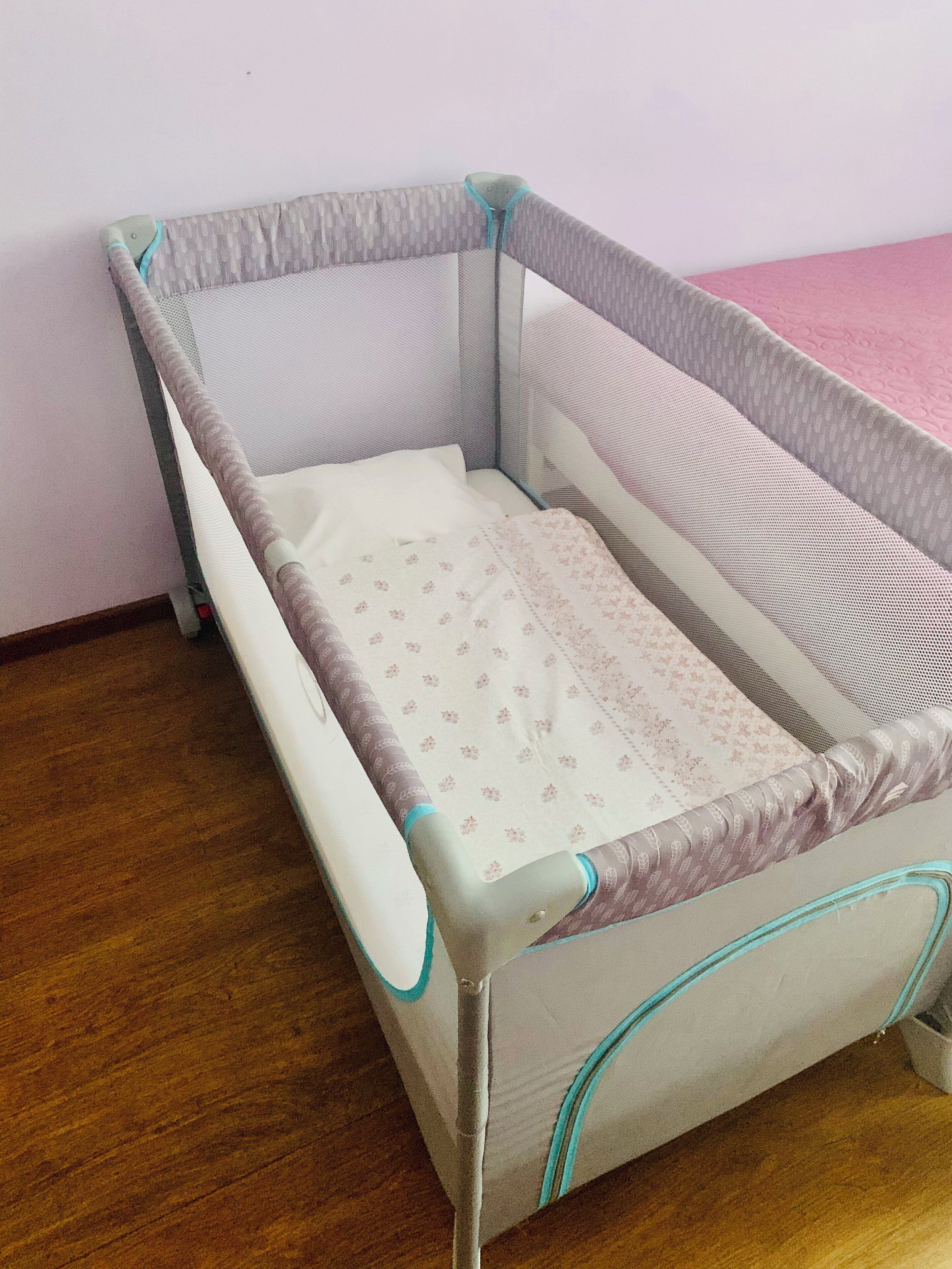 A baby crib with a mesh side and a padded mattress, placed against a wall with a pink surface.