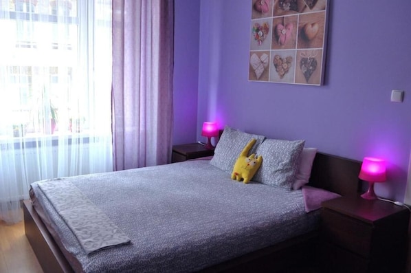 A bedroom with a bed, two bedside lamps, a window with curtains, and a wall with a heart-themed artwork. - Happy House Hostel. Poznan (Poznan)