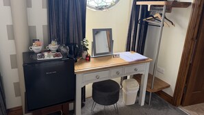 Een kleine kamer met een bureau, een minibar, een spiegel, een prullenbak en een raam met gordijnen.