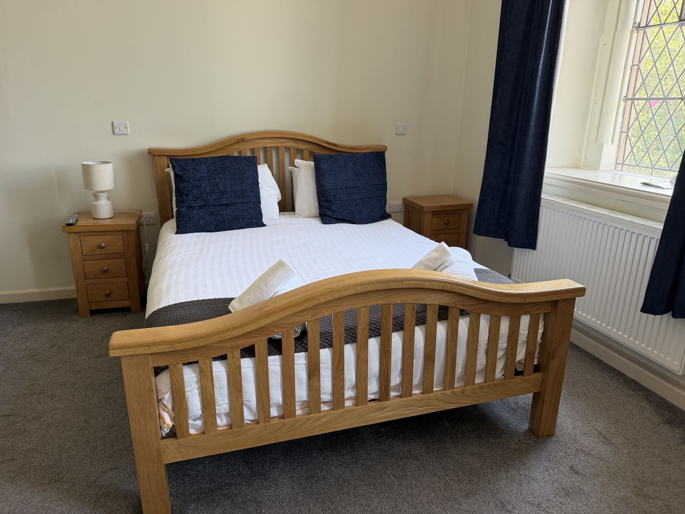 A bedroom with a wooden bed, white bedding, a bedside table with a lamp, and a window with curtains.