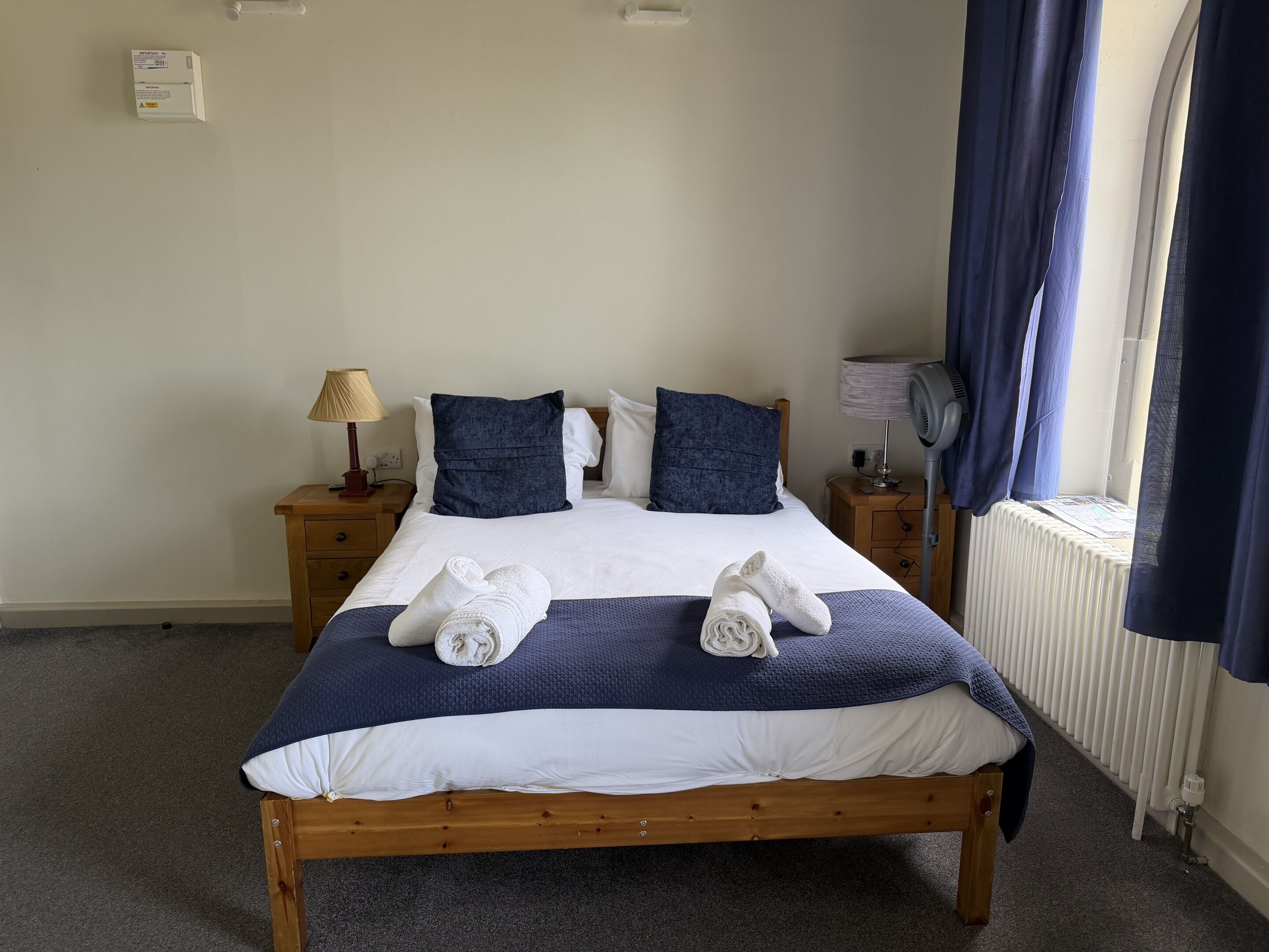 A neatly made bed with two towels, a wooden headboard, a nightstand with a lamp, and a window with curtains.
