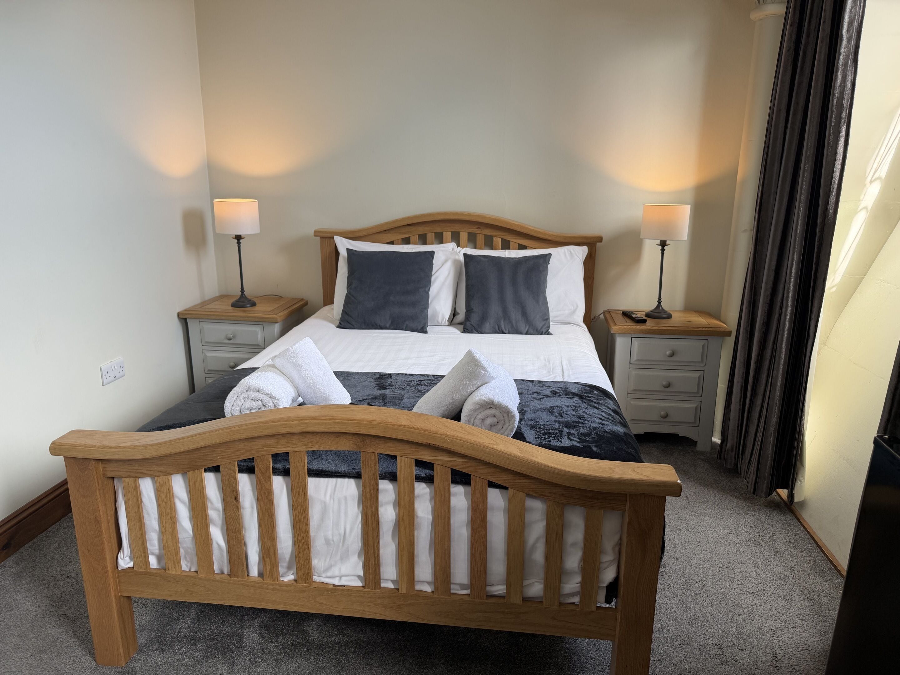 Un lit bien fait, avec une tête de lit en bois, deux tables de chevet avec des lampes et un rideau foncé d’un côté.