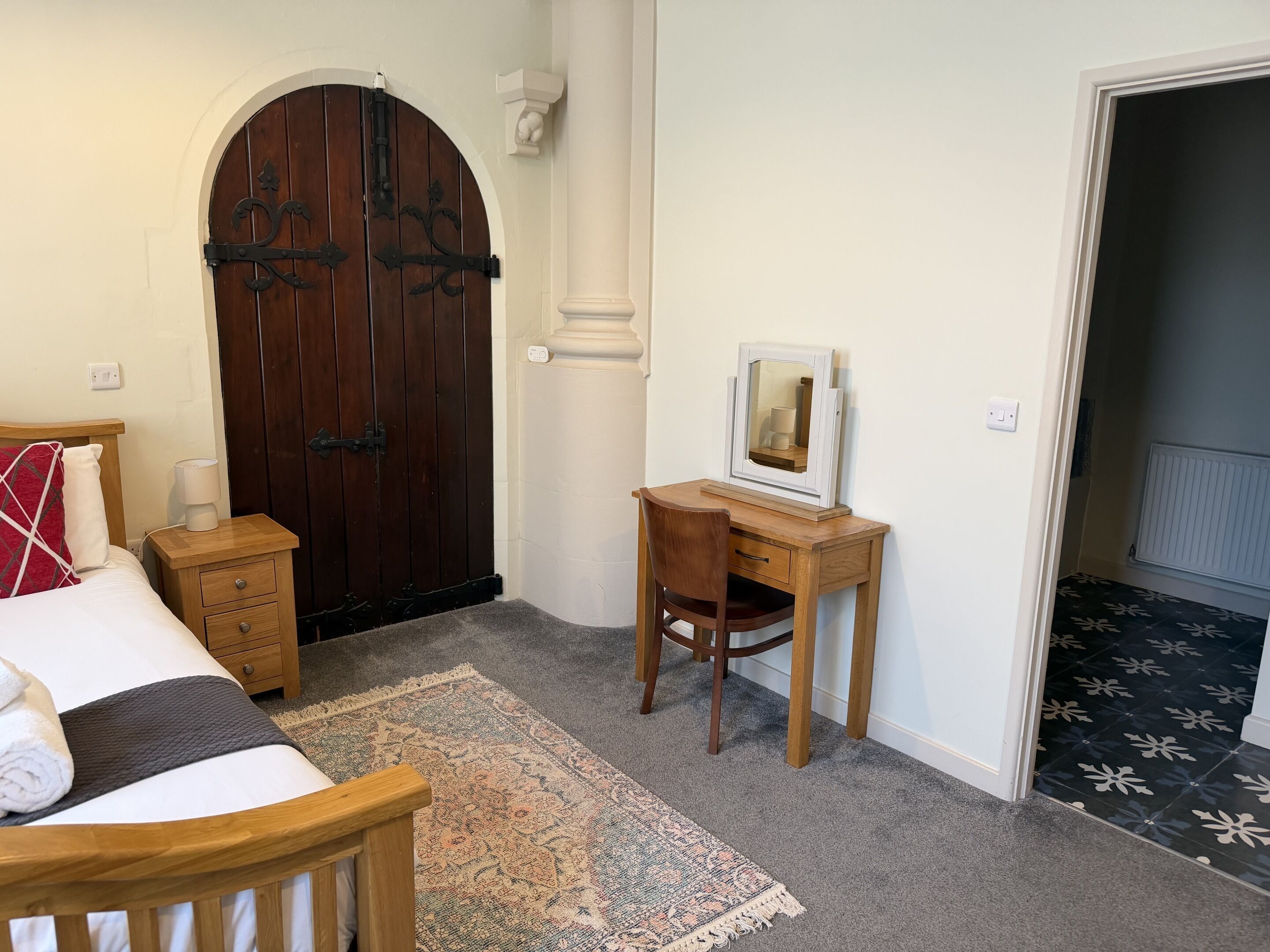 A bedroom with a bed, a desk, a chair, a rug, and a large wooden door.