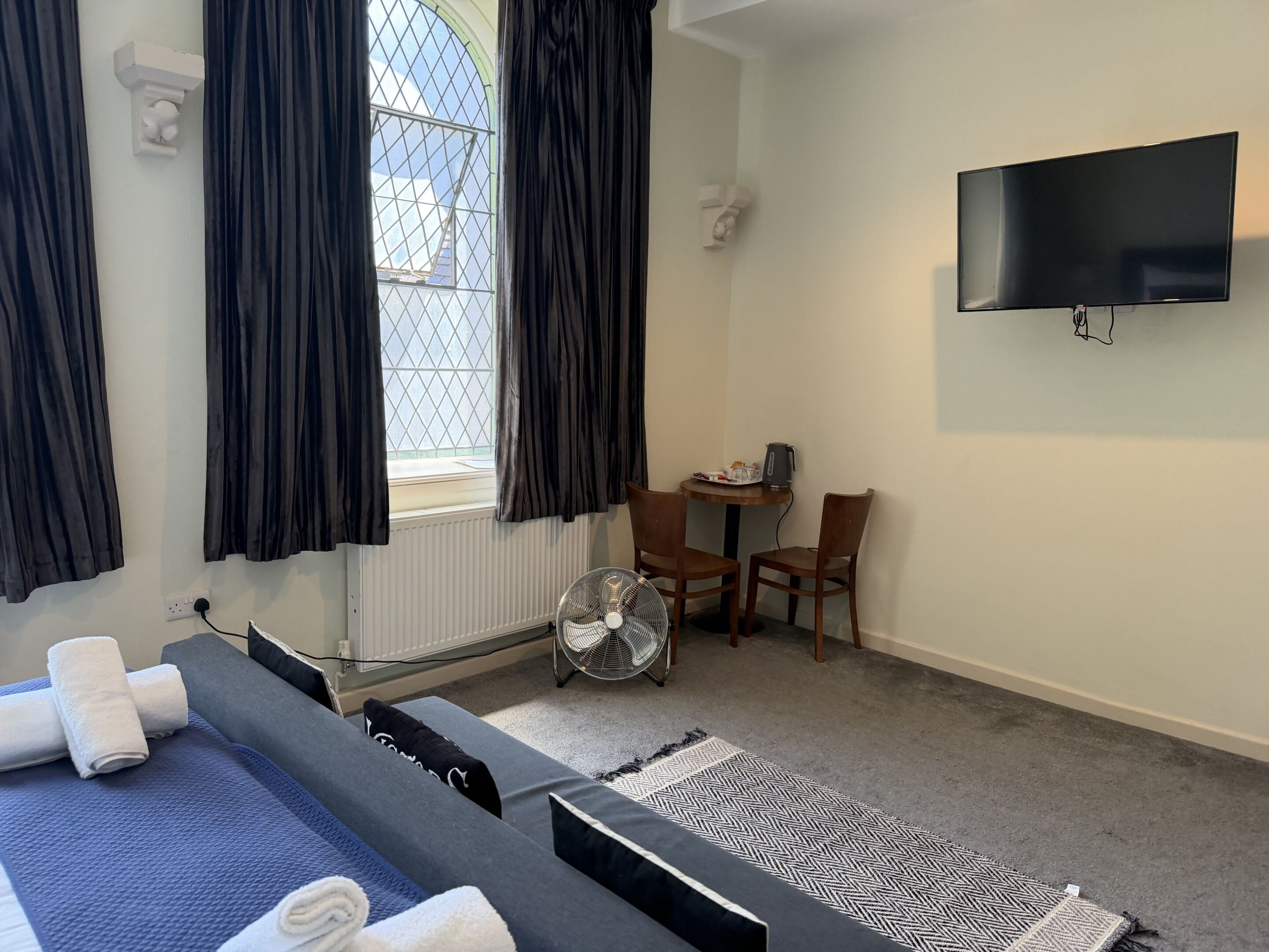 A hotel room with a bed, a desk, a chair, a TV, a window with curtains, and a radiator.