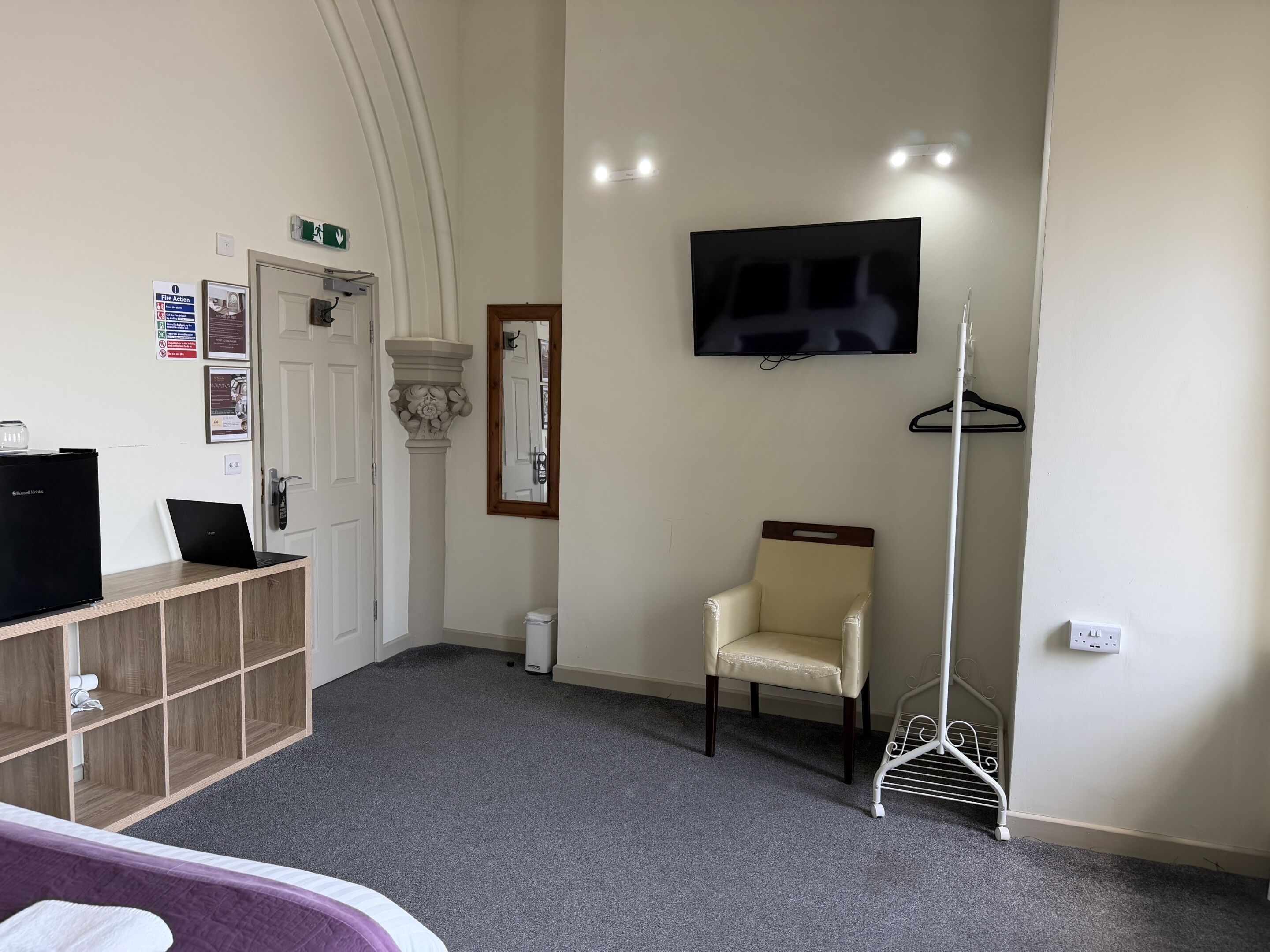 A hotel room with a bed, a desk with a computer, a chair, a TV, and a coat rack.