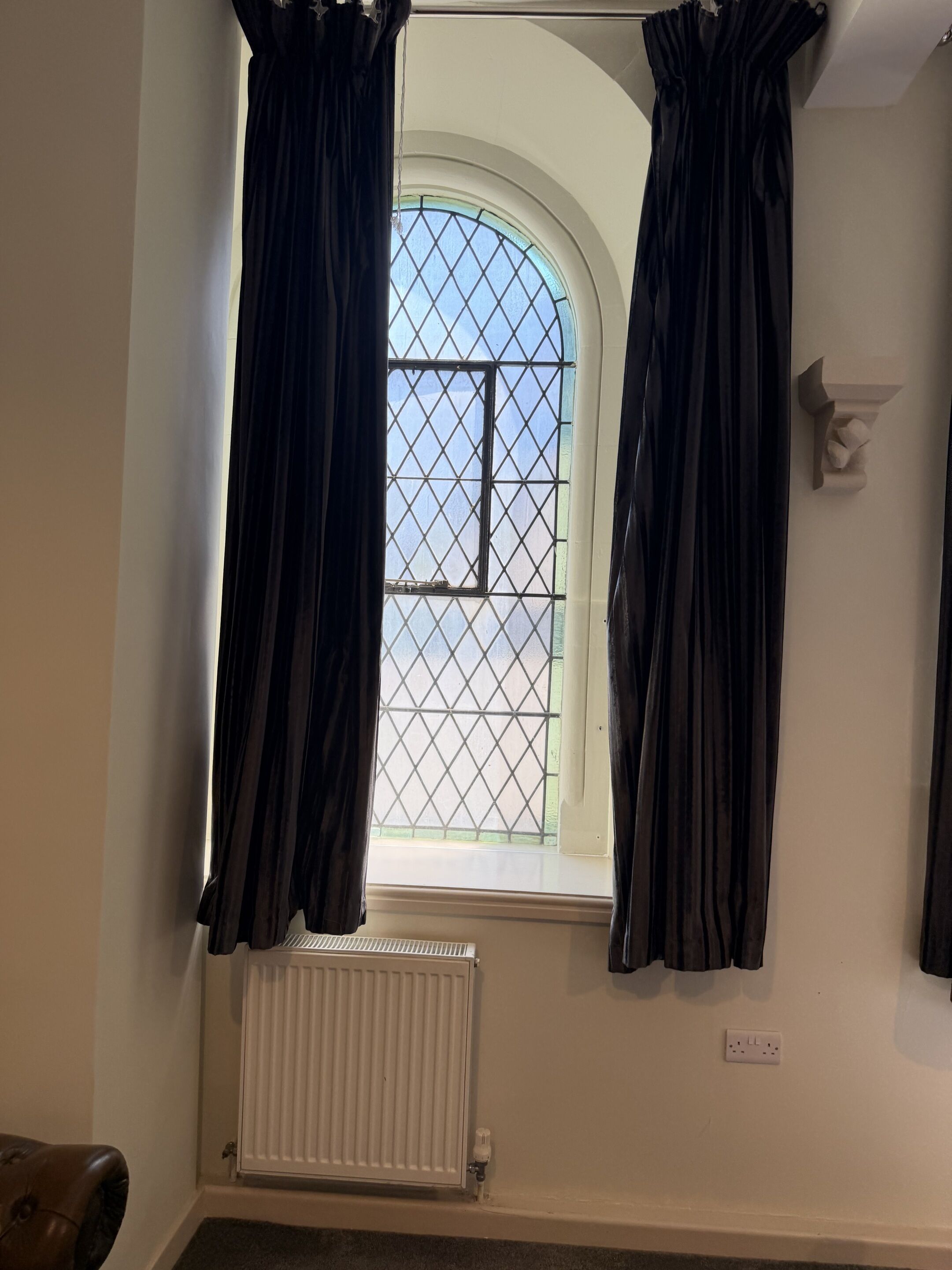 A room with a window, curtains, a radiator, and a wall-mounted light fixture.