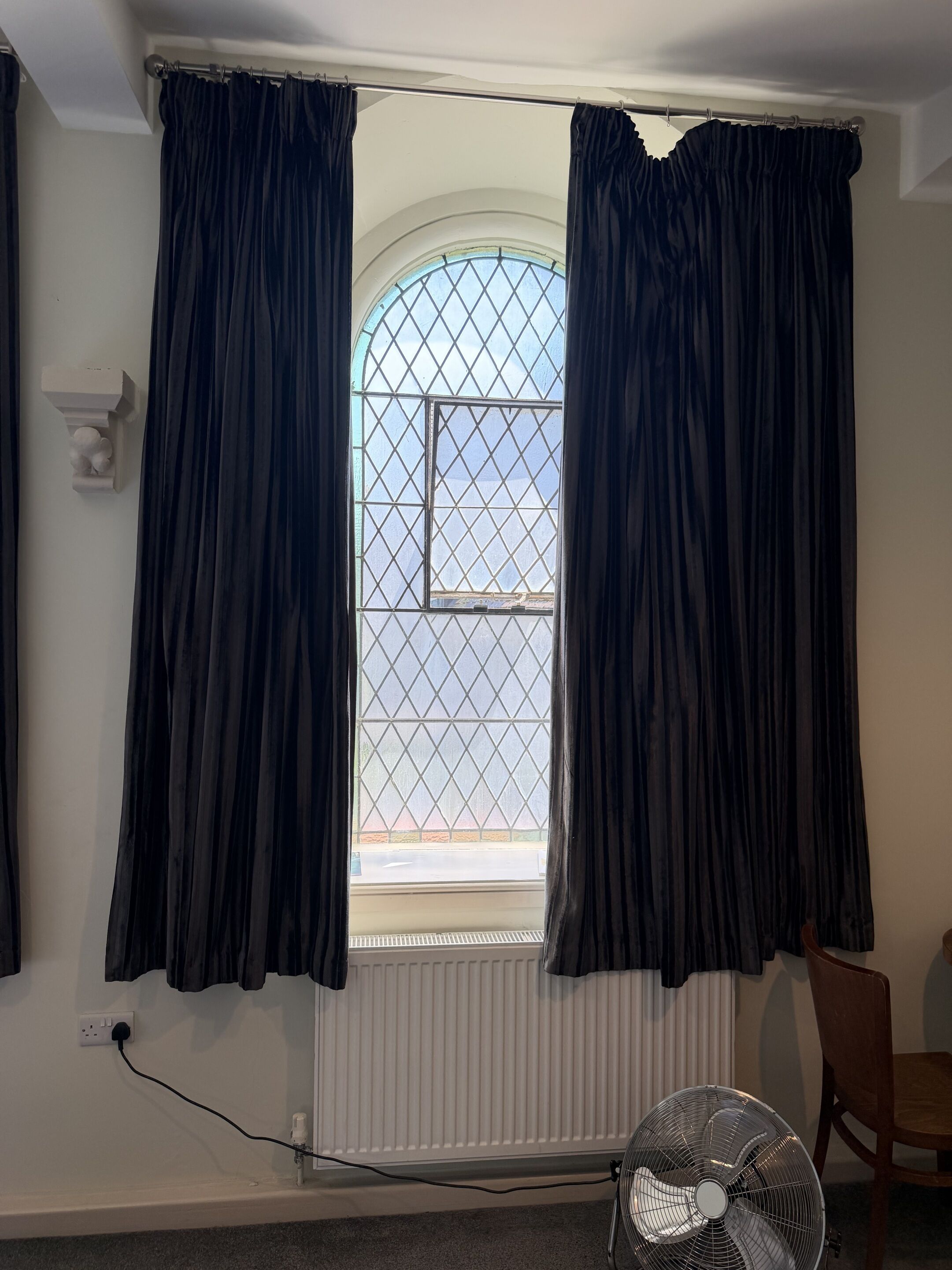 A room with a window covered by dark curtains, a radiator, a power outlet, and a standing fan.