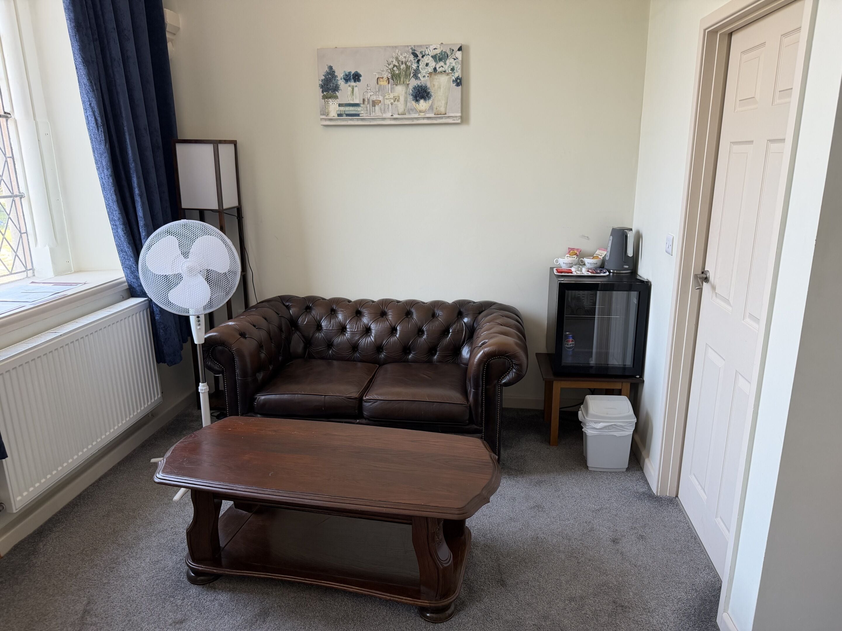 A living room with a leather sofa, a wooden coffee table, a fan, a painting, a cabinet, and a door.
