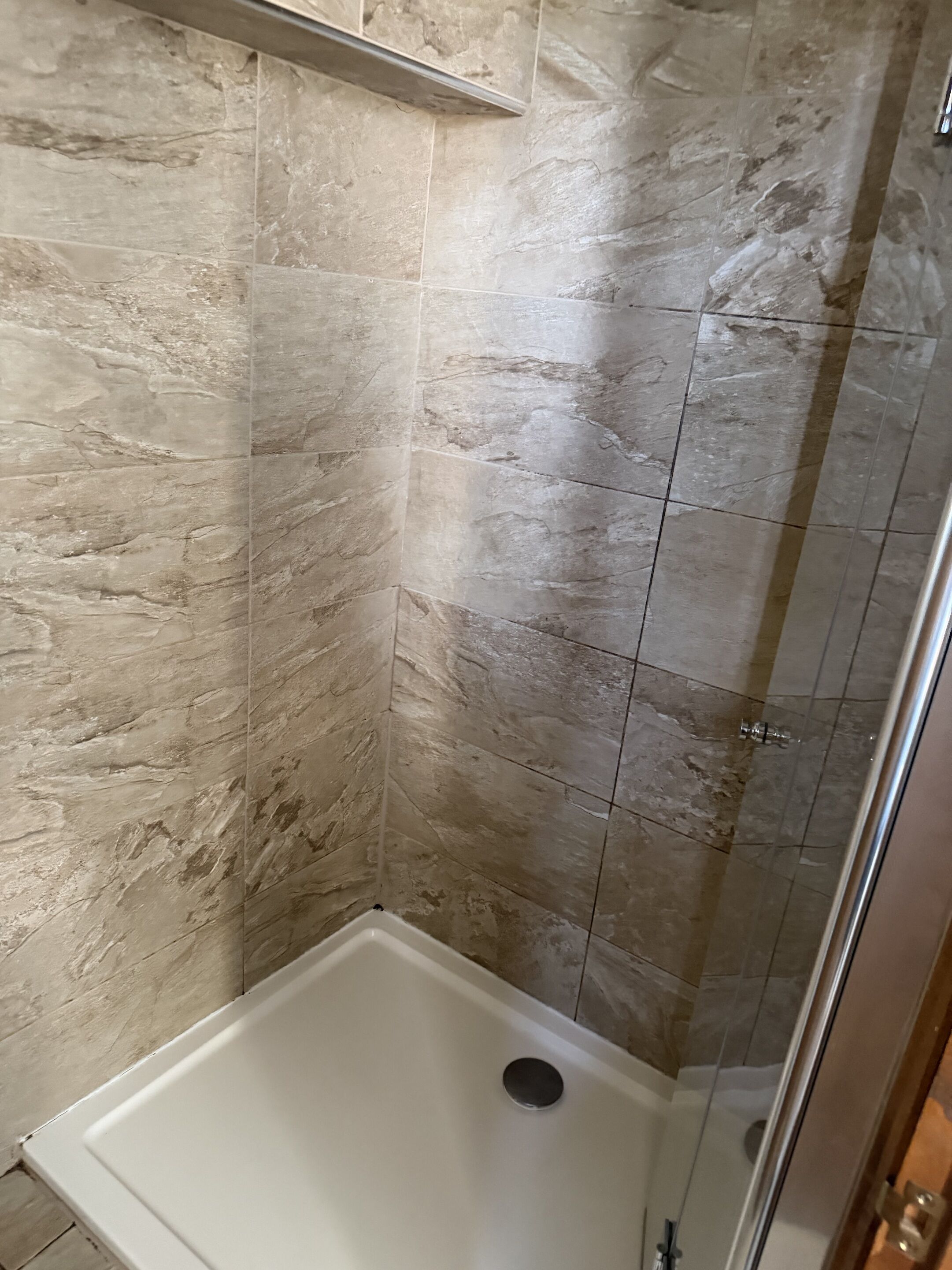 A shower area with a glass door, beige marble tiles, and a white bathtub.