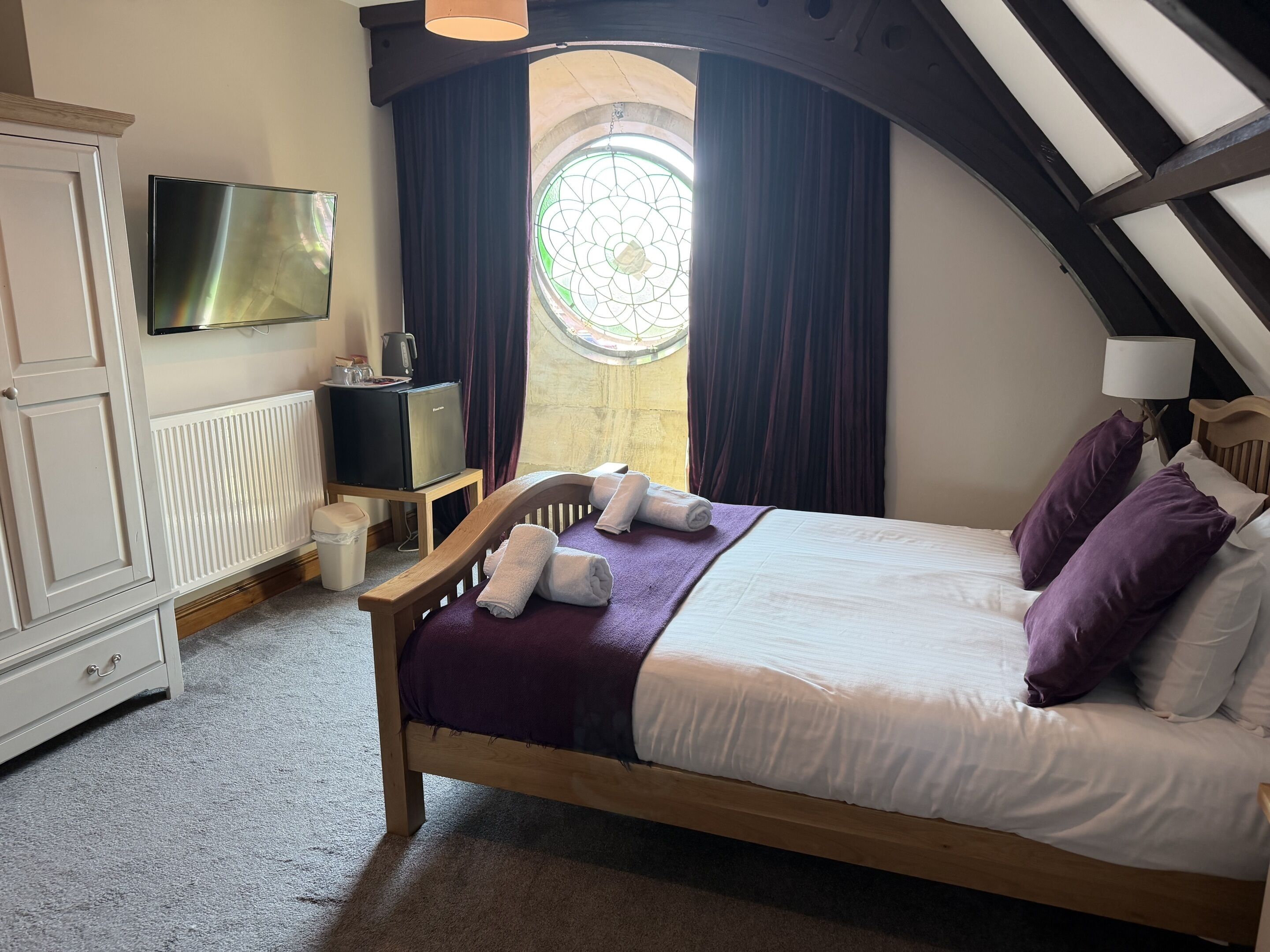 A bedroom with a bed, a television, a radiator, a window with curtains, and a wooden ceiling.