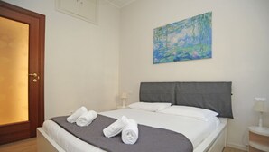A neatly made bed with white linens and a gray headboard, a painting of a water scene on the wall, and a wooden door. - Gambara Apartment Travel Inside (Milano)