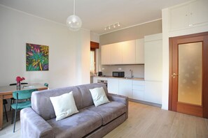 A modern living room with a grey sofa, a dining table with chairs, a kitchen area with white cabinets, and a wooden door with a frosted glass panel.