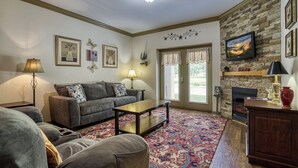 A living room with a sofa, coffee table, and a fireplace. There are framed pictures on the wall and a television mounted above the fireplace. - Fantastic and Convenient Smoky Mountain View Condo! 2104 (Pigeon Forge)
