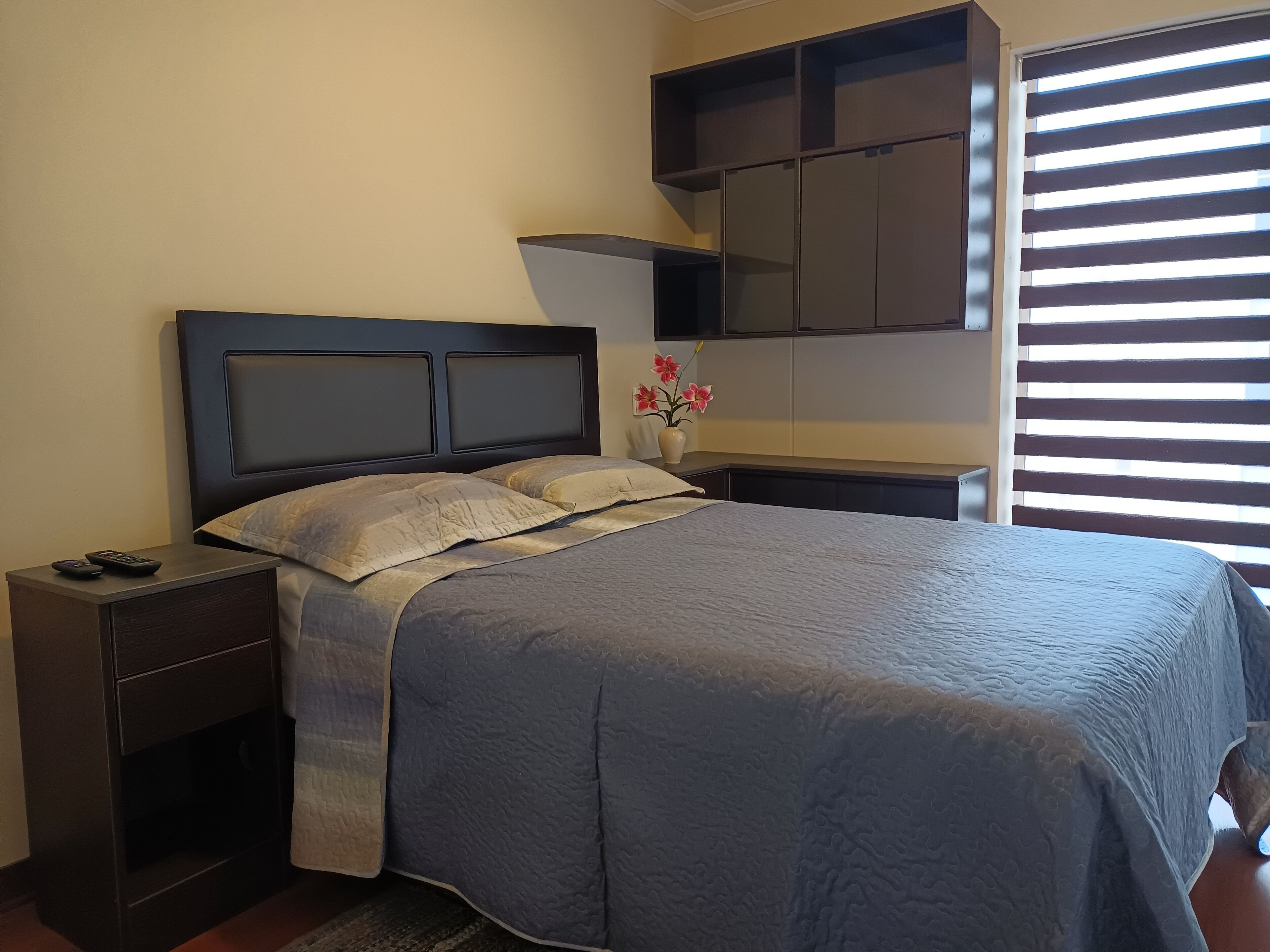 A bedroom with a bed, nightstands, a dresser, a window with blinds, and a wall-mounted cabinet.