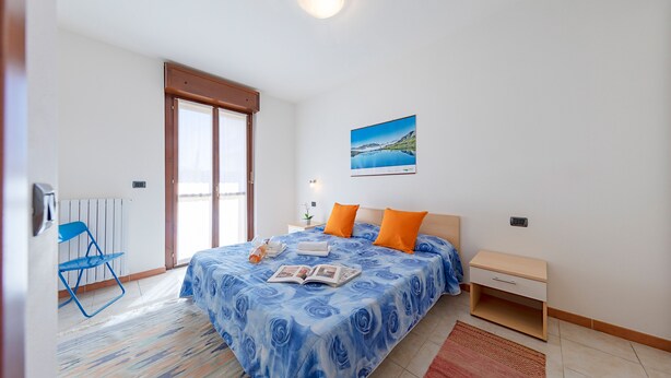 A bedroom with a bed, a nightstand, a chair, a window, and a painting on the wall. - La Mimosa B03 Apartment by Wonderful Italy (Peschiera del Garda)