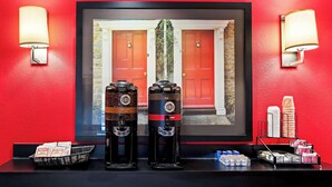 Two coffee machines, a basket of magazines, and a stack of disposable cups on a counter with a red wall and framed window in the background. - Suburban Studios Columbus Fort Benning Area (Columbus)