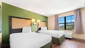 A hotel room with two beds, a large window with curtains, a wall-mounted air conditioner, and a headboard with wall sconces. - Suburban Studios Columbus Fort Benning Area (Columbus)
