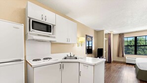 A compact kitchen with a microwave, stove, and sink. It features white cabinetry and a small window with curtains. - Suburban Studios Columbus Fort Benning Area (Columbus)