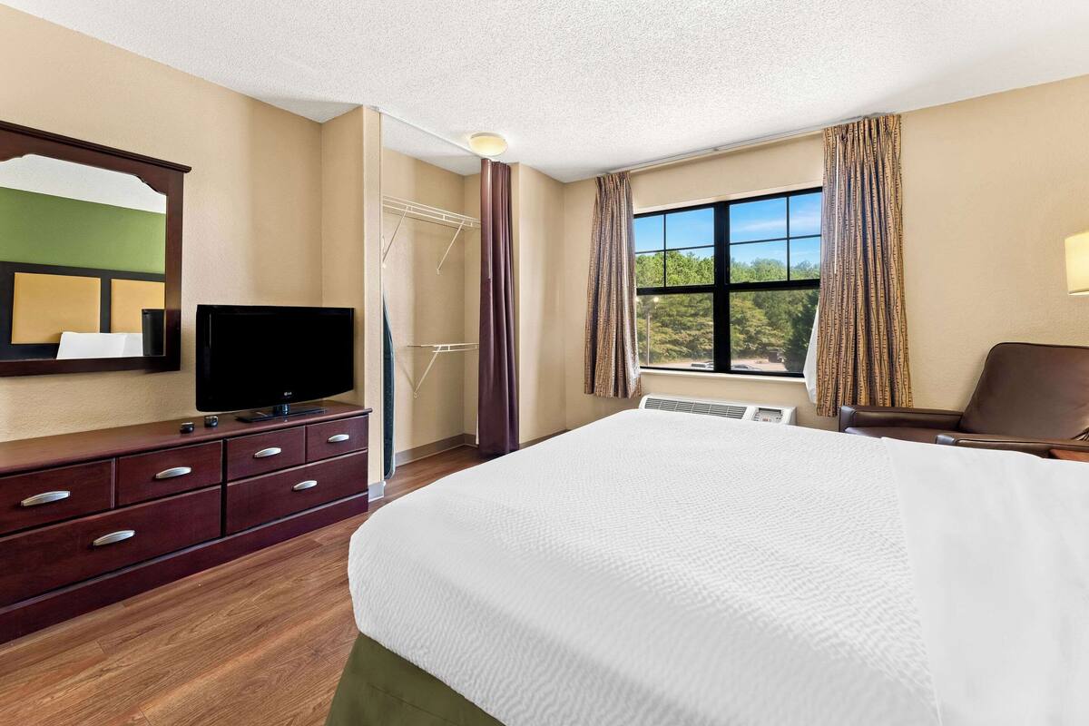 Une chambre d’hôtel avec un grand lit, un téléviseur posé sur une commode en bois, une fenêtre avec des rideaux et une vue sur les arbres.