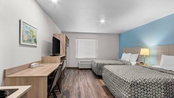 Habitación de hotel con escritorio, televisor, cama con ropa de cama estampada, lámpara de noche y ventana con persianas.