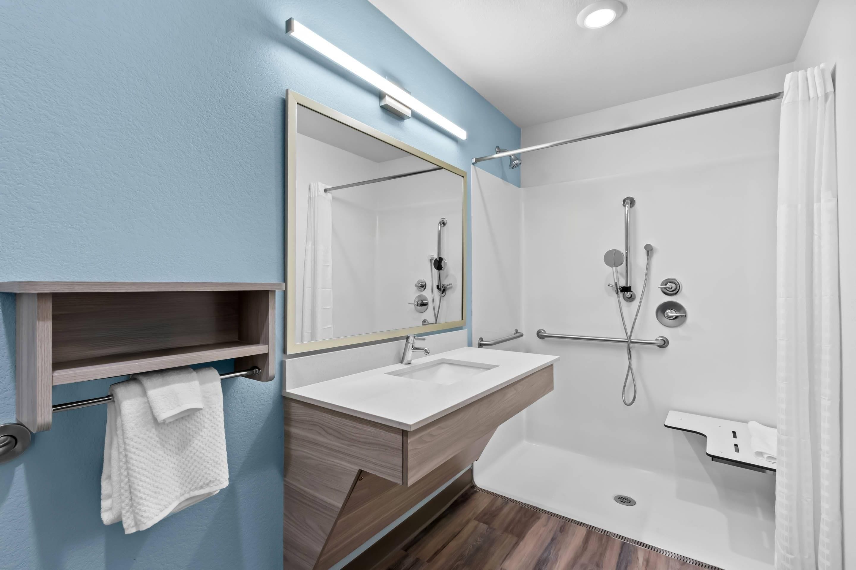 A modern bathroom with a wooden vanity, a large mirror, a shower with a handheld showerhead, and a towel rack.