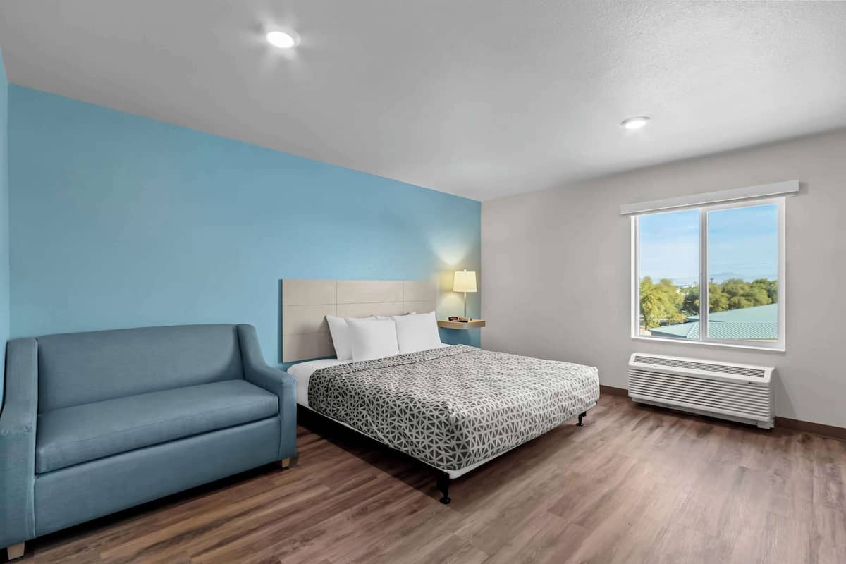 A bedroom with a bed, a blue sofa, a nightstand with a lamp, a window with a view, and a wall-mounted air conditioner.