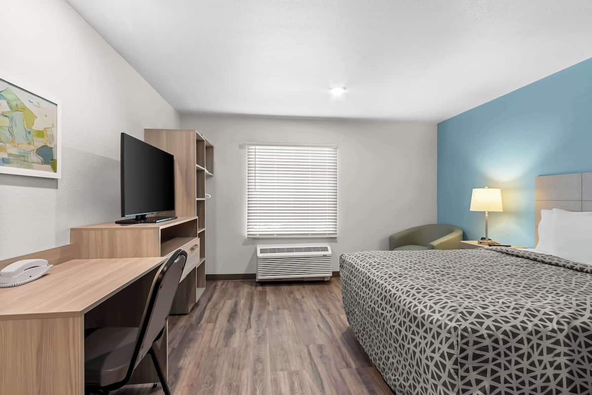 A hotel room with a desk, chair, TV, air conditioning unit, bed, and a lamp.