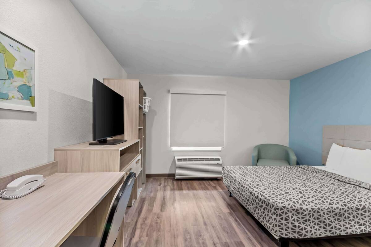 A hotel room with a bed, a desk with a telephone, a television, a window, and a wall-mounted air conditioner.