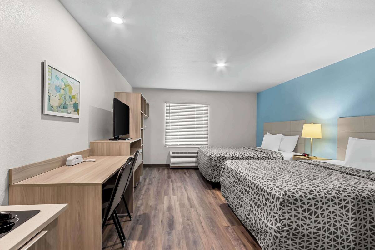 A hotel room with a desk, two beds, a TV, a lamp, and a painting on the wall.
