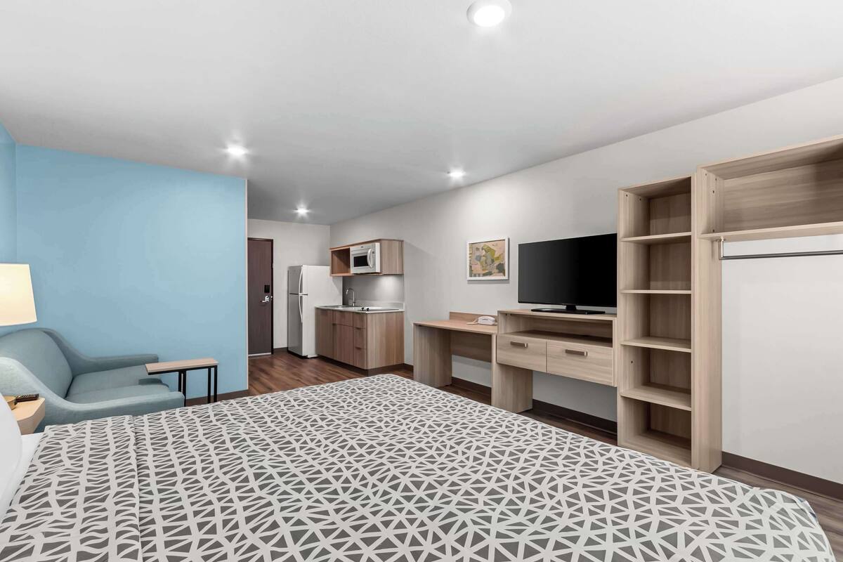 A modern hotel room with a bed, a desk, a TV, a kitchenette, and a wardrobe.