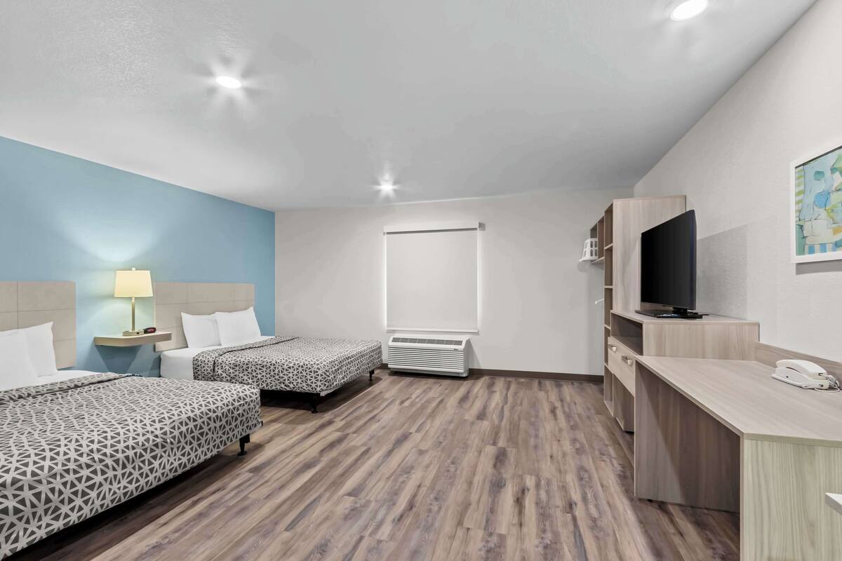 A hotel room with two beds, a desk, a TV, and a wall-mounted air conditioner.