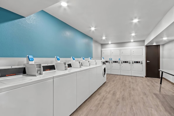 A commercial laundry facility with multiple washing machines and dryers, a blue accent wall, and a wooden floor. - Woodspring Suites Gilbert - Mesa Gateway Airport (Gilbert)