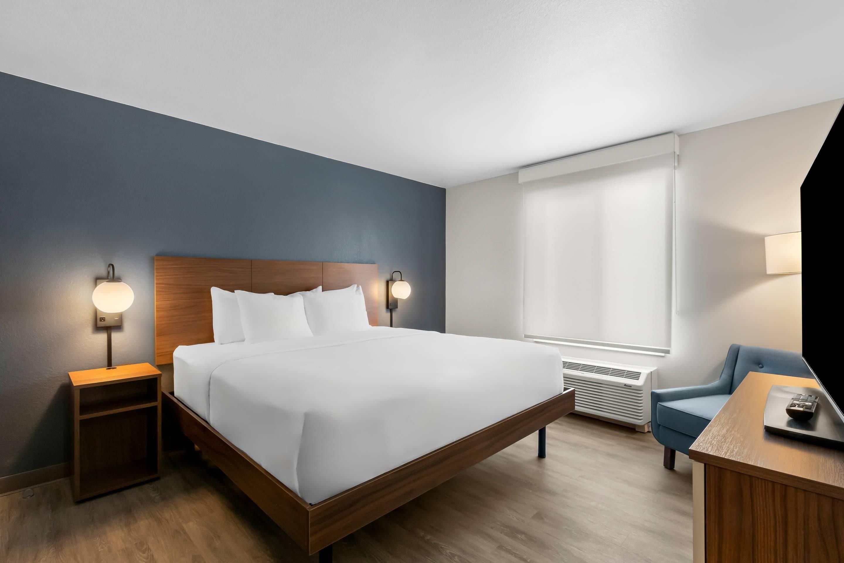 Habitación de hotel con cama de madera, lámparas de noche, ventana con persianas, una silla azul y un escritorio con lámpara.