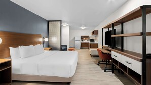 A modern hotel room with a large bed, a desk, a TV, and a kitchenette in the background.