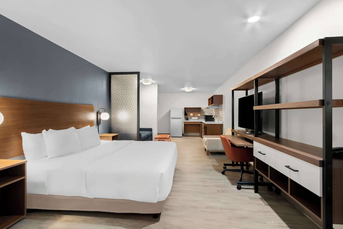 Una habitación de hotel moderna con una cama grande, un escritorio, un televisor y una kitchenette al fondo.