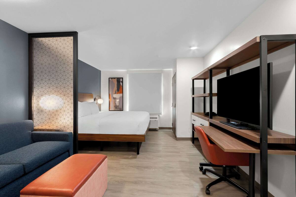 Una habitación de hotel moderna con cama, escritorio con computadora, televisión, sofá y una otomana naranja pequeña.