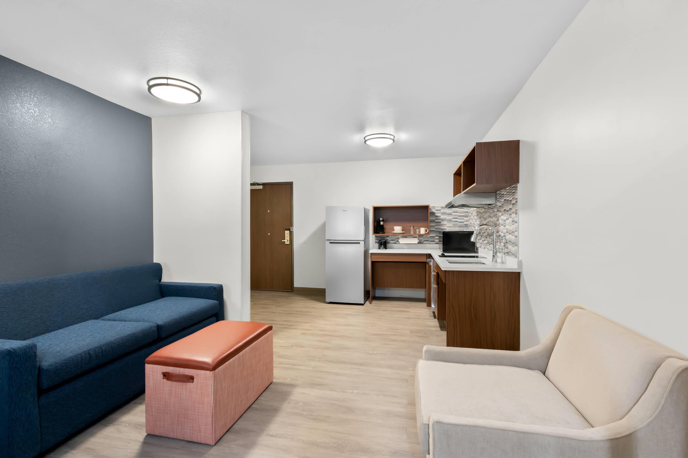 A modern living room with a sofa, ottoman, and a kitchenette in the background.