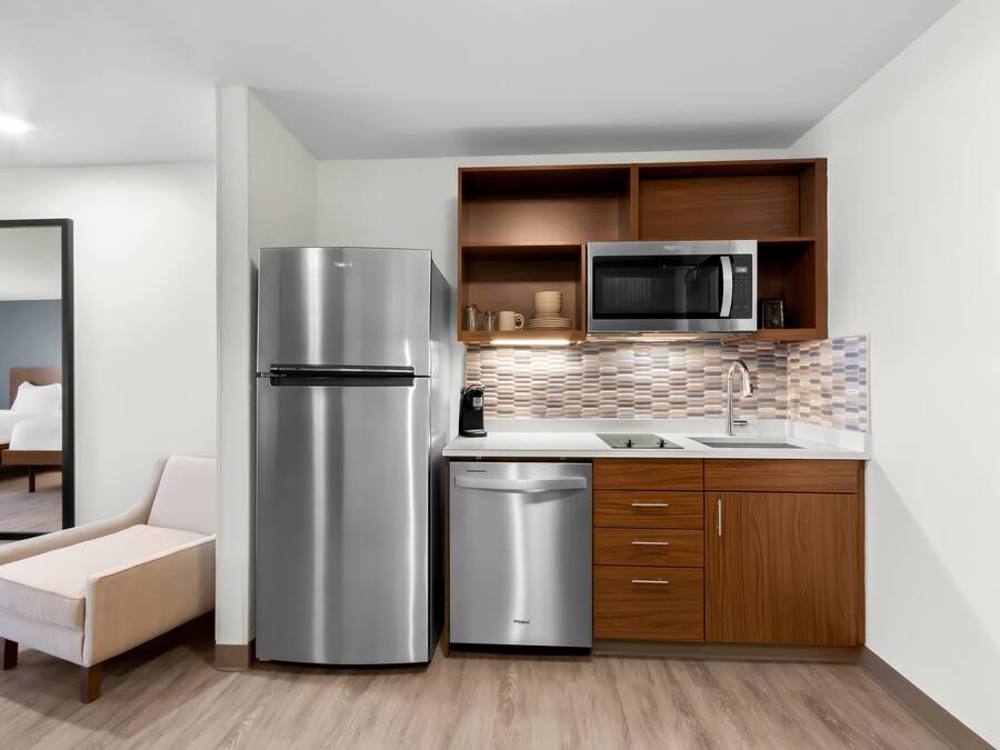 A compact kitchen with a stainless steel refrigerator, a microwave, and a dishwasher. There is a wooden cabinet above the sink and a small seating area with a chair.