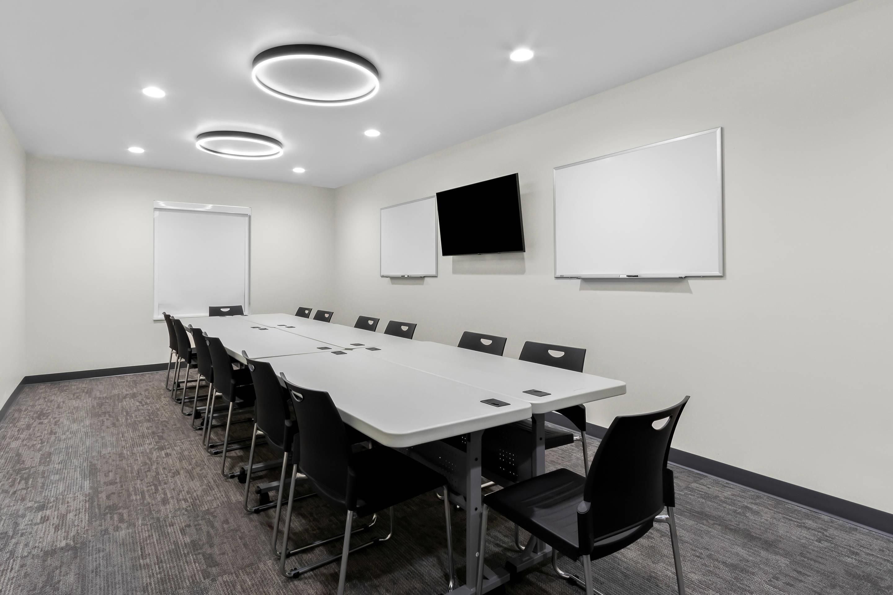 A conference room with a long table, black chairs, a whiteboard, a flat-screen TV, and recessed ceiling lights.
