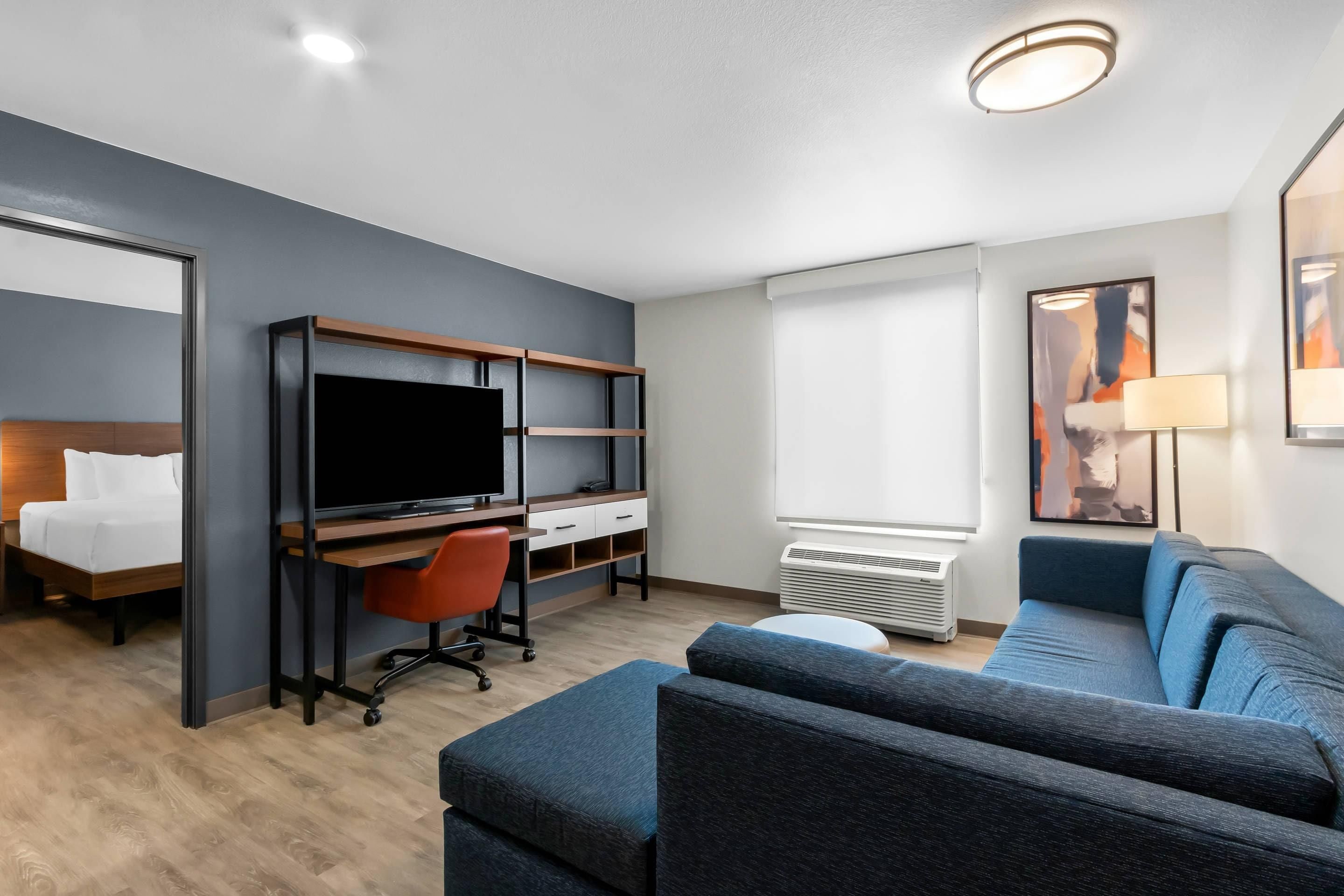 A modern hotel room with a bed, a desk with a computer, a sofa, and a large window.