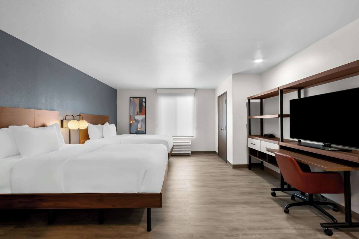 Habitación de hotel con una cama grande, un escritorio con computadora, un televisor y una ventana con persianas.