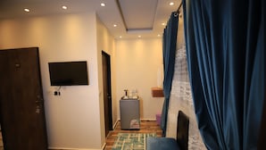 Ein Hotelgang mit Holztür, Flachbildfernseher, Minibar, gemustertem Teppich und blauem Vorhang.