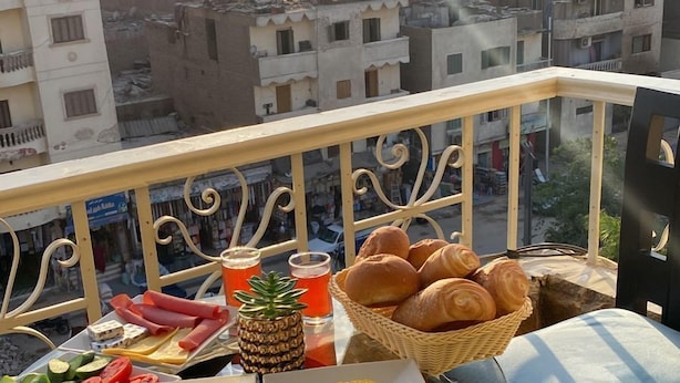 Ein Balkon mit einem Tisch, gedeckt für das Frühstück, darunter Croissants, Tomaten, Käse und eine Ananas. Im Hintergrund sind Pyramiden sichtbar.