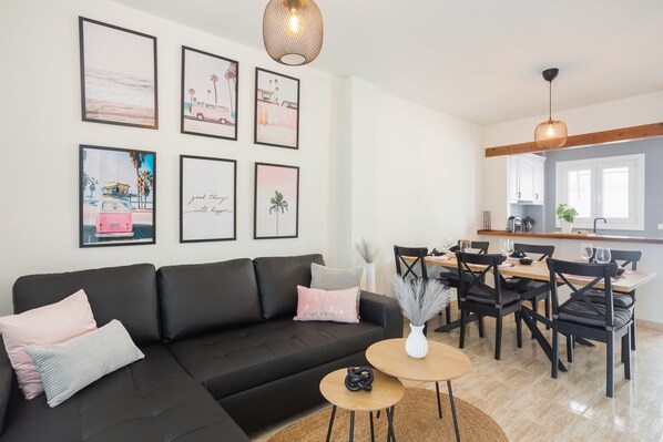 A modern living room with a black sofa, a dining area with a wooden table and chairs, and a kitchen in the background.