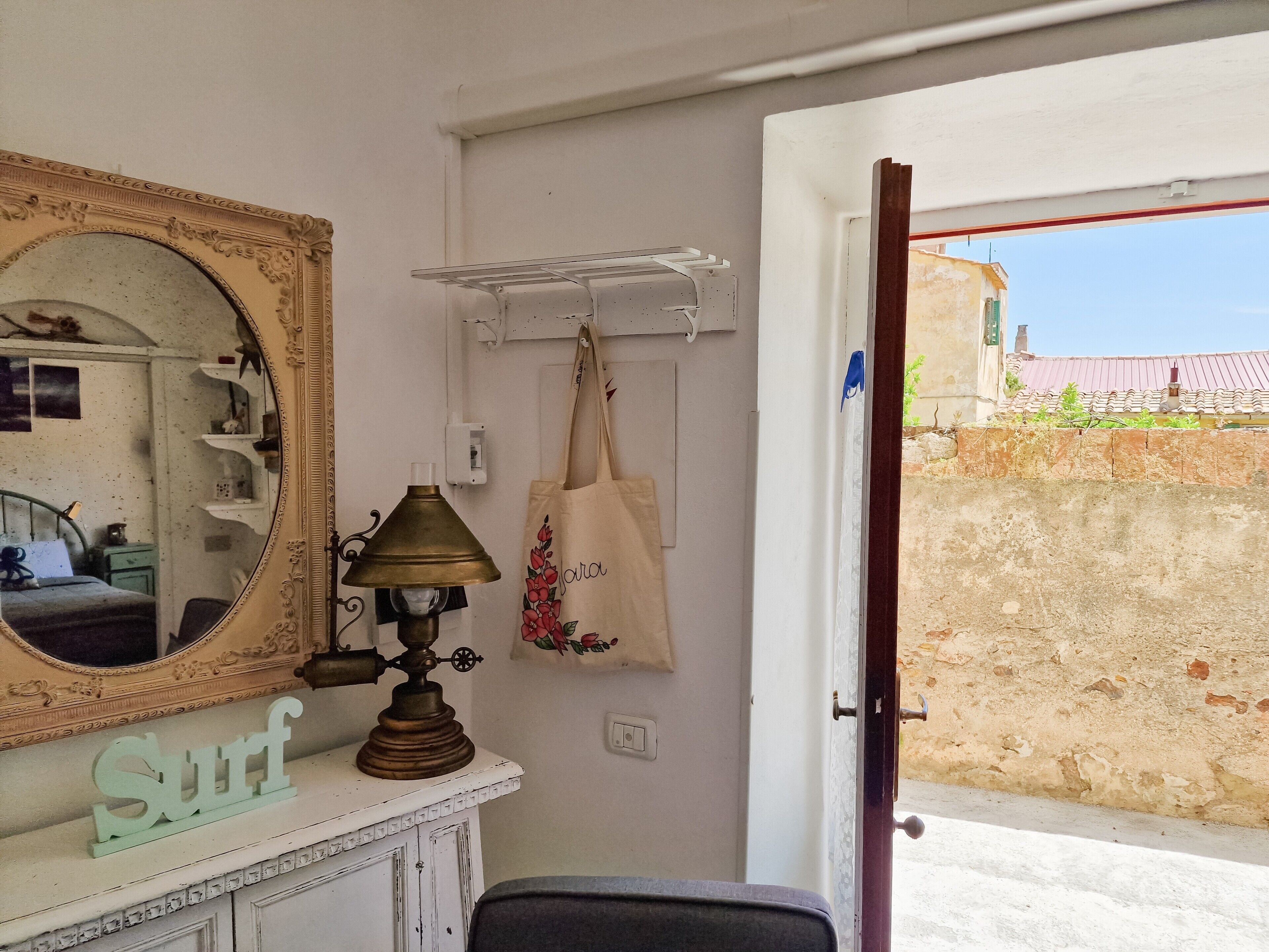 A room with a mirror, a vintage lamp, a surf-themed decoration, a hanging bag, and a door leading to an outdoor area.