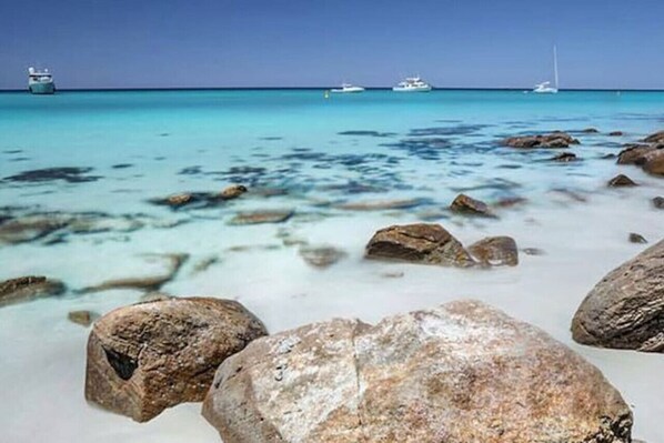 Una spiaggia con acque turchesi cristalline, grandi rocce in primo piano e imbarcazioni all'orizzonte.