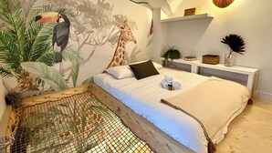 Een slaapkamer met een muurschildering van een toekan en een giraffe, een bed met witte lakens, een klein plankje met decoratieve objecten, en een hangende lamp.
