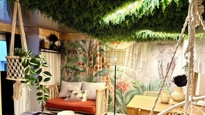 Een knus vertrek met een hangstoel, een houten tafel, een bank met kussens, en een muurschildering met tropische planten.