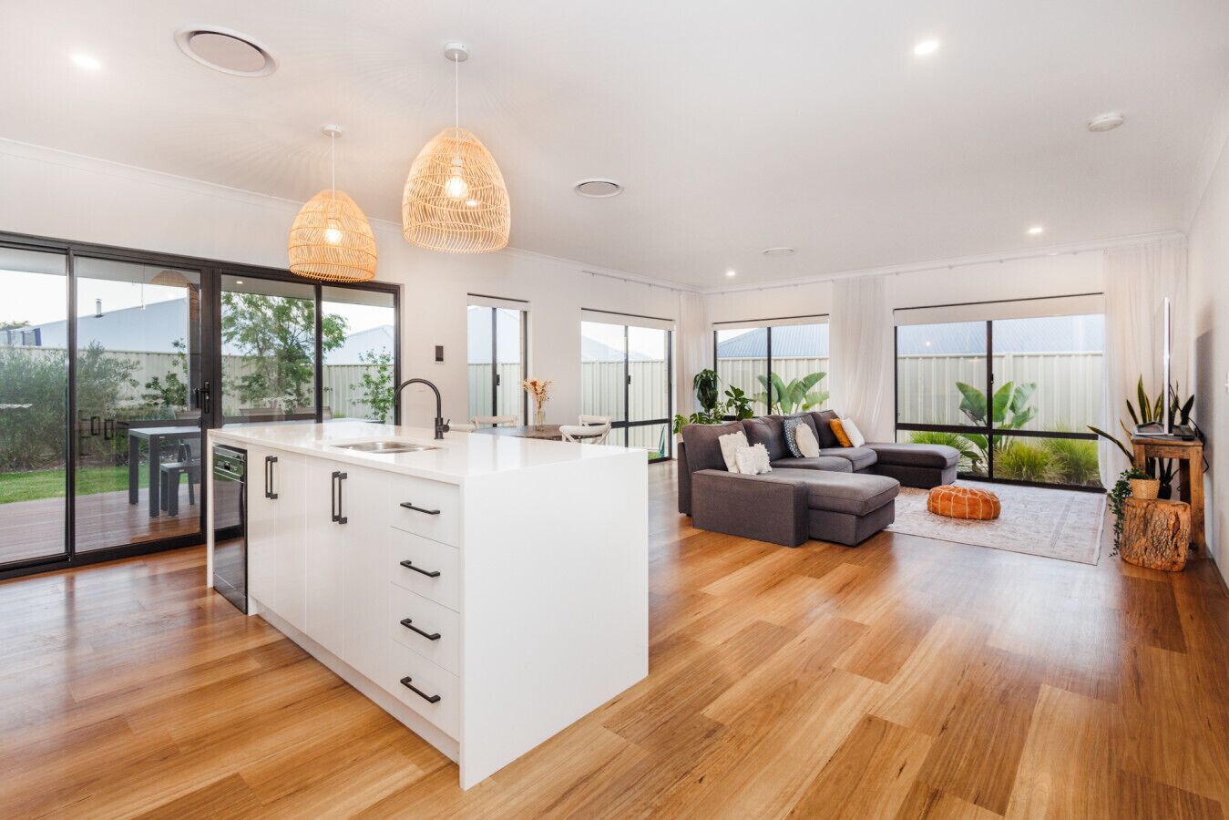 A modern kitchen with a large island, a dining area with a table and chairs, a gray sofa, and large windows with a view of greenery.